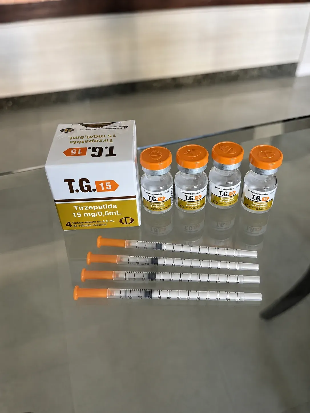 Kit da TG 15 Tirzepatida 15 mg/0,5 mL para revenda: caixa original com faixa dourada, quatro frascos com tampa laranja e seringas, sobre superfície refletiva